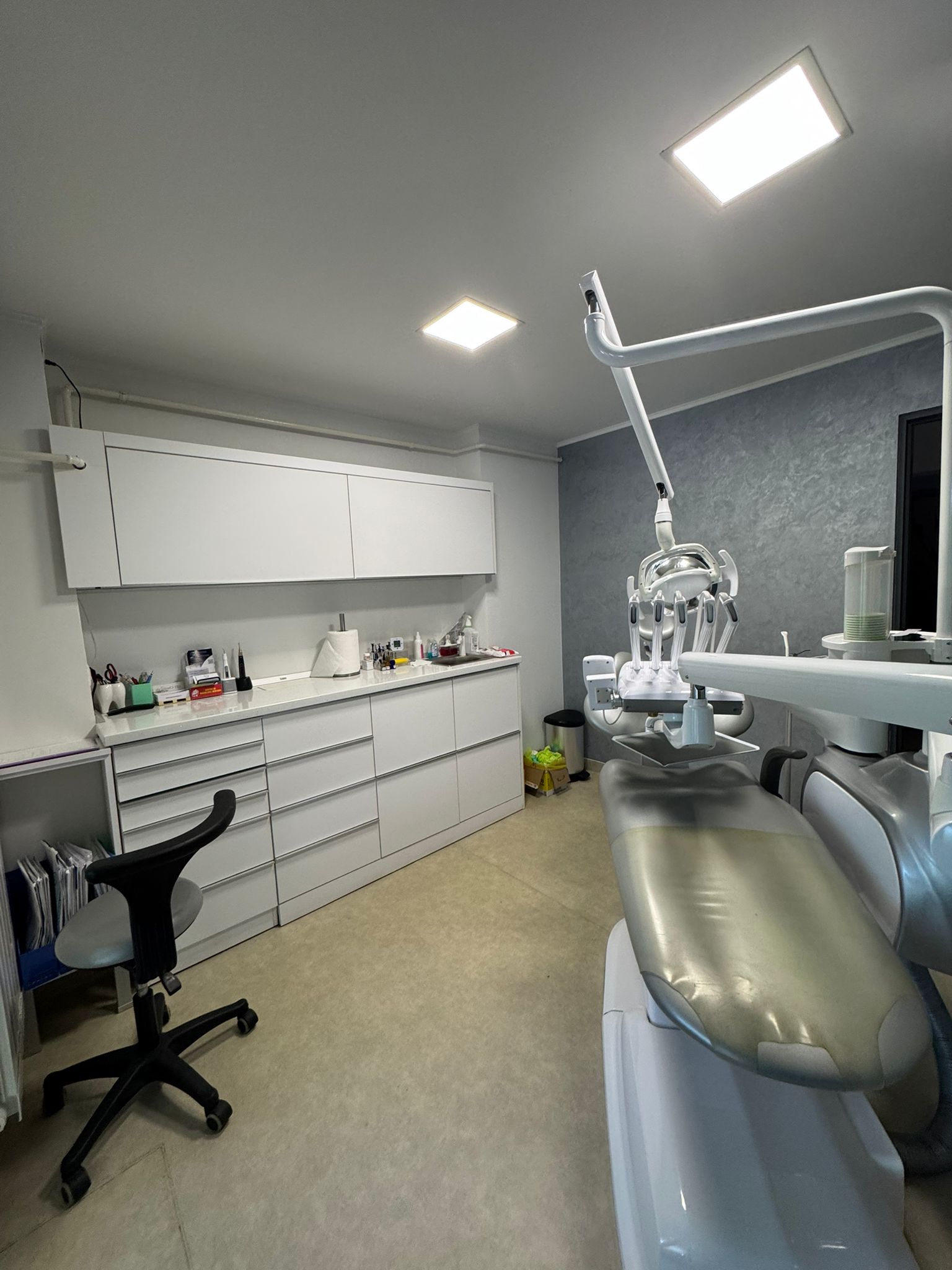 Medic dentist tânăr îmbrăcat în halat albastru, cu brațele încrucișate, în cabinet stomatologic cu scaun verde și echipamente dentare albe.
